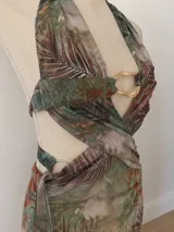 Vestido midi con estampado botánico en tonos tierra y verdes, con escote halter profundo y detalle de argolla metálica dorada en el centro. Presenta aberturas laterales en la cintura y falda con volados laterales.