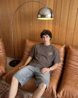 Remera de corte oversize color marrón oscuro, con estampa gráfica circular en color crema en la espalda que presenta tipografía estilizada y detalles de texto en la parte inferior.