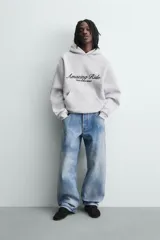 Sudadera con capucha y bolsillo frontal tipo canguro, confeccionada en tejido de algodón con interior perchado. Presenta diseño de corte boxy fit, mangas largas con puños y ruedo en rib, y texto bordado y estampado en el frente.