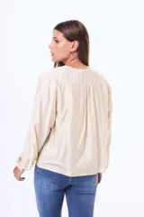 Blusa de satén color beige, con cuello redondo, detalle de volados en los hombros y cierre frontal con lazo. Presenta mangas largas con puños elásticos.