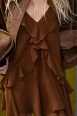 Vestido corto color marrón con escote en pico y tirantes anchos. Presenta un diseño con múltiples volantes en cascada que recorren el frente y el bajo, con un acabado asimétrico y forro interior a tono.