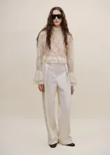 Blusa de encaje color crudo, con cuello alto, mangas largas con puños acampanados y volados en el bajo.