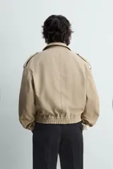 Cazadora color beige, estilo cropped, con cuello de solapa, manga larga con puños ajustables y trabillas en los hombros. Presenta bolsillos de vivo en la cadera y cierre frontal de botonadura.