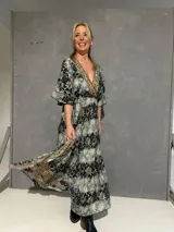 Vestido largo cruzado al frente de seda estampado en tonos azules, negros y dorados, con escote en V ribeteado, mangas 3/4 fluidas y elástico en la parte trasera de la cintura.
