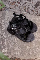 Sandalias negras de estilo chunky con tiras cruzadas en charol y base de goma dentada. Cuentan con una tira superior desmontable y cierre con hebilla metálica en el tobillo.