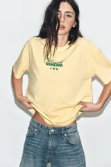 Camiseta amarilla de corte oversize, con cuello redondo, manga corta y estampa frontal en color verde con texto alusivo a Brasil Ipanema y pequeños símbolos.