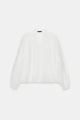 Camisa de manga larga en color blanco, con cuello redondo, escote en pico y cierre frontal con botones. Presenta detalles de volantes en el mismo tejido a lo largo de la abertura frontal.