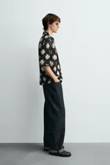 Camisa de manga corta con cuello solapa y cierre frontal de botones, confeccionada en tejido de crochet con patrón de flores en negro y beige.