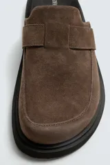 Zueco marrón de piel con acabado serraje, detalle de cinta en el empeine, plantilla acolchada, horma redonda y suela con volumen.