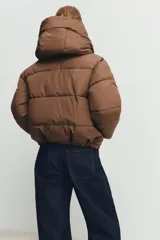 Campera puffer acolchada de color negro, con cuello envolvente, capucha integrada, manga larga, bolsillos laterales ocultos y bajo con elástico. Cierre frontal con botones a presión ocultos por solapa.