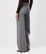 Pantalón de jean color gris lavado, con corte wide leg de piernas amplias y cintura baja. Presenta detalle de tachas metálicas a lo largo de las costuras laterales, cierre frontal con botón y bolsillos funcionales.