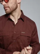 Camisa de manga larga color marrón, con cuello clásico, cierre frontal con botones y dos bolsillos frontales con solapa en el pecho.
