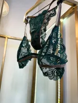 Conjunto de lencería compuesto por un bralette de encaje verde esmeralda con cierre frontal y una tanga a juego con tiras regulables, ambos con detalles de ribetes en color marrón.