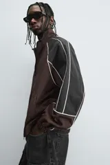 Cazadora de ajuste relajado, color marrón oscuro con bloques de color negro en las mangas. Presenta ribetes blancos que delinean los paneles y cuello alto con cierre frontal de cremallera. Los puños y el bajo son elásticos.