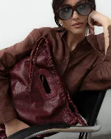 Cartera tipo tote de cuero color bordó, con diseño fruncido en los laterales y abertura central integrada como asa. Cuenta con una silueta flexible y espaciosa, ideal para uso diario.