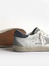 Zapatillas Golden Goose modelo Super-Star de cuero blanco con estrella de gamuza gris, refuerzo de talón con purpurina gris oscuro, aplicación trasera de gamuza gris y cordones color crema.