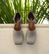 Zapato de cuero vacuno color gris topo, con talón descubierto y pulsera con hebilla. Tiene taco de madera forrado en cuero y suela de goma. La horma es de punta cuadrada, ancha y cómoda.