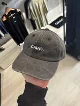 Gorra con visera de efecto acid wash en color gris oscuro, con diseño de seis paneles y el texto 'GAINS.' bordado en blanco en el frente.