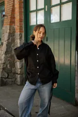 Camisa de lino color negro con silueta relajada, cuello camisero y cierre frontal con botones de nácar. Presenta mangas largas con puños y detalles de tiras laterales.