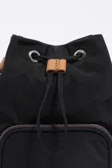 Bolso tipo bucket de Bimba y Lola, confeccionado en nylon negro con detalles en cuero color suela. Cuenta con cierre de cordón ajustable, bolsillo frontal con cierre y correa de hombro regulable de tejido.