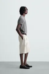 Bermuda color beige claro, de corte ancho y largo, confeccionada en algodón. Presenta cierre frontal con cremallera y botón, bolsillos laterales, bolsillo cerillero frontal y bolsillos traseros de vivo.