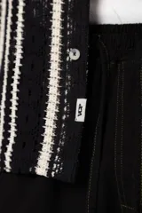 Camisa de manga corta con cuello clásico y cierre frontal de botones. Confeccionada en tejido de punto crochet calado, presenta un patrón de rayas verticales negras sobre fondo crudo.