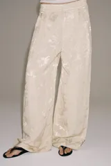 Pantalón de tiro medio confeccionado en hilatura con lino, con cintura con trabillas, bolsillos laterales ocultos y pierna ancha. Presenta un diseño con estampado jacquard de motivos florales en tonos beige y crudo, y cierre frontal con cremallera, botón y ganchos metálicos.