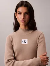 Vestido mini de Calvin Klein color beige, confeccionado en tejido de punto acanalado con cuello alto, manga larga y ajuste ceñido al cuerpo. Cuenta con una etiqueta con el logo de la marca en el pecho.
