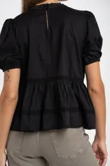 Blusa negra de algodón con corte oversize, mangas cortas abullonadas y detalles de encaje y bordados en el frente y hombros. Presenta un diseño con pliegues verticales y terminaciones con puntilla.