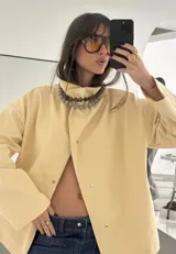 Campera de algodón color amarillo manteca, con cuello alto, cierre frontal con botones metálicos y cinturón ajustable en la cintura. Presenta un diseño versátil que permite llevarla entallada o con silueta holgada.