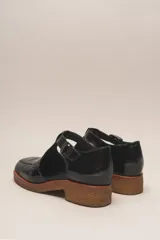 Zapato tipo guillermina de cuero charol color marrón oscuro, con tira T y hebilla ajustable en el lateral. Posee puntera cuadrada y suela de goma color caramelo con taco bajo.