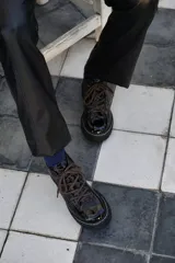 Borcegos de cuero charolado negro con cordones jaspeados en tonos marrones y suela track dentada con costura blanca visible.