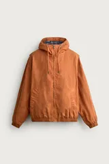 Cazadora de corte relajado para hombre, confeccionada en algodón con acabado efecto encerado en color marrón. Cuenta con cuello subido, capucha ajustable con cordones, mangas largas con puños elásticos y cierre frontal con cremallera. Incluye bolsillos frontales de vivo y forro interior a cuadros.