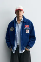 Cazadora estilo varsity confeccionada en pana de algodón azul, con interior ligeramente acolchado. Presenta cuello solapa, cierre frontal con botones a presión, bolsillos laterales de vivo y acabados en rib a rayas en puños y cintura. Incluye múltiples parches bordados decorativos en el frente.