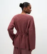 Cardigan de tricot en color borravino, con textura acanalada, escote en V profundo y cierre frontal con un único botón metalizado. Presenta mangas largas y un diseño alargado con amarre ajustable en la espalda.