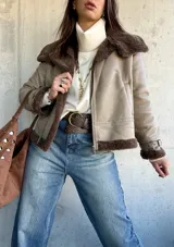 Campera tipo gamulán de cuero sintético en color vizón, con interior y solapas de piel sintética en color marrón oscuro. Presenta cierre frontal, correas ajustables con hebillas en los puños y diseño de corte recto.