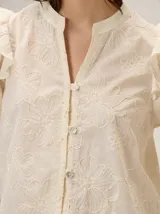Camisa de color crema con bordados florales, cuello mao, cierre frontal con botones y mangas largas abullonadas con volados en los hombros.
