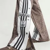 Pantalón Adidas Adibreak de corte holgado, confeccionado en poliéster reciclado con estampado animal print tipo jirafa en tonos marrón y blanco. Cuenta con bandas laterales blancas con líneas negras y botones a presión, además del logo clásico de la marca.
