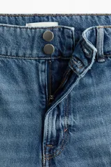 Pantalón de jean de tiro alto con corte wide leg, confeccionado en denim de algodón rígido. Presenta cierre frontal con doble botón, pasacintos y pliegues marcados en la parte delantera y trasera.