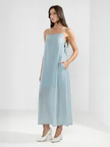 Vestido largo de color celeste confeccionado en tejido arrugado liviano. Presenta un diseño con escote recto, breteles finos y detalles de lazos en los laterales. Incluye bolsillos laterales, forro interno corto y un ruedo asimétrico con caída posterior más larga.