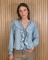 Camisa de denim claro, con cuello en V adornado con volados grandes a lo largo de la botonadura frontal, mangas largas con puños fruncidos elastizados y botones oscuros.