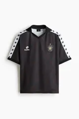 Camiseta de futbol de manga corta con corte holgado, color negro con estampa sutil y bandas blancas con rombos en las mangas. Presenta cuello en V con solapa y esc11udo bordado en el pecho.