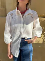 Camisa blanca de corte amplio con mangas tres cuartos abullonadas. Presenta detalles de bordado calado en los hombros y en las mangas, con cierre frontal de botones.