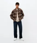 Camisa tipo overshirt de corte oversized, confeccionada en tejido de poliéster con textura. Presenta un estampado de cuadros en tonos marrones y beige, cuello clásico, cierre frontal con botones y bolsillo aplicado en el pecho.