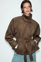 Chaqueta de gamuza sintética color marrón, con cuello subido, cierre frontal cruzado con botones y mangas largas anchas. Cuenta con bolsillos delanteros y cinturón ajustable del mismo tejido.