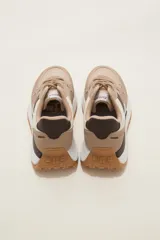 Zapatilla deportiva de diseño urbano, combinando paneles de tela blanca y gamuza sintética en tono gris claro. Cuenta con cordones grises, suela de plataforma con entresuela blanca y base dentada en color caramelo.