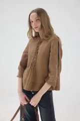 Blusa de lino color marrón, de corte recto y mangas 3/4. Presenta escote redondo con pequeños volados y pliegues frontales, además de detalles de encaje en las mangas y puños con volados.