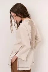 Chaqueta beige de lino con mangas 3/4, cuello mao y detalles de costuras decorativas oscuras en contraste.