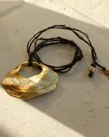 Collar artesanal con dije de bronce macizo en forma orgánica irregular, similar a una concha marina, suspendido de un cordón de seda natural negro. El diseño permite diversas formas de uso, ya sea ajustado al cuello o largo.