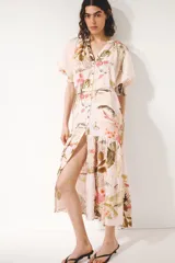 Vestido midi oversized de mezcla de viscosa, con estampado floreado tropical sobre base rosada, escote en V y botonadura frontal. Cuenta con mangas al codo raglán con puños fruncidos, cinturón fino para atar con cuentas y diseño escalonado en la falda.
